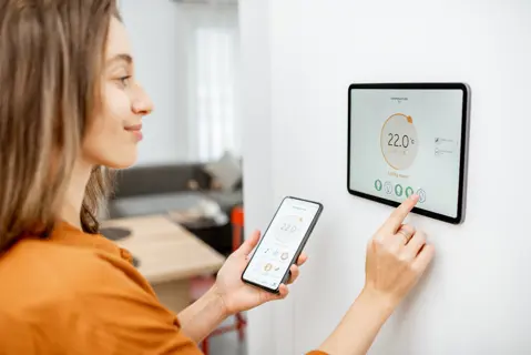 Eine Frau steuert die Temperatur im Wohnzimmer mit Smartphone und digitalem Touchscreen-Panel.